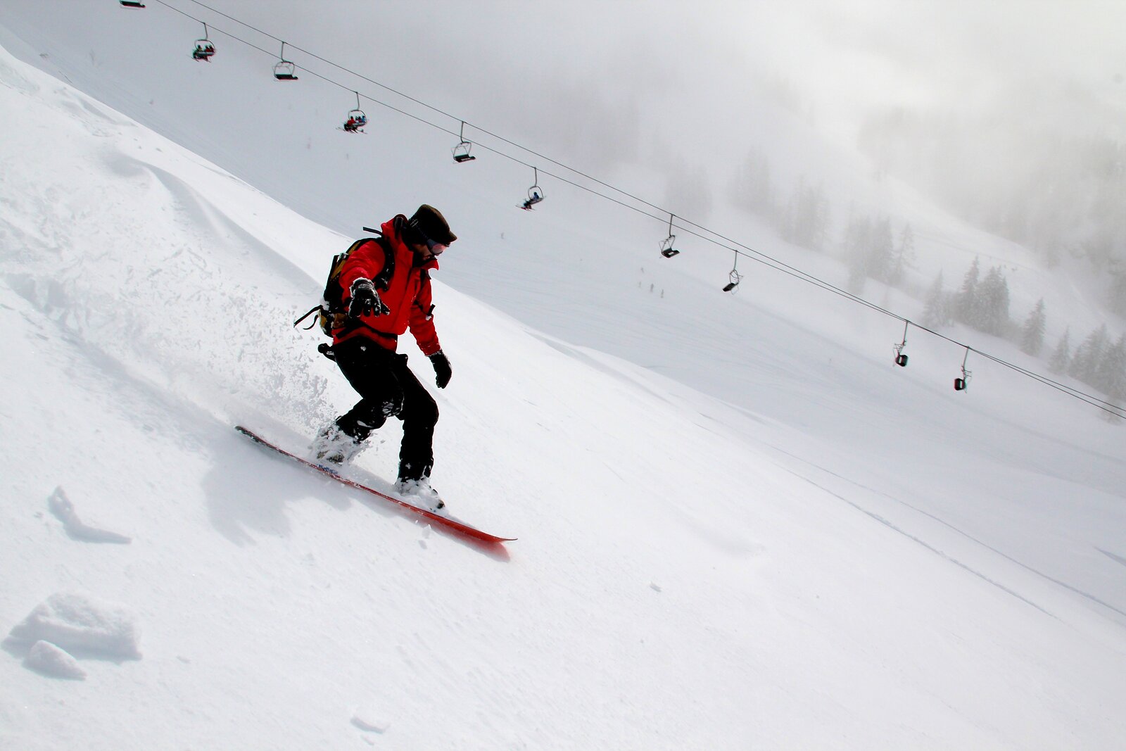 Snowboardista v červené bundě sjíždí zasněžený svah pod sedačkovou lanovkou v mlze.