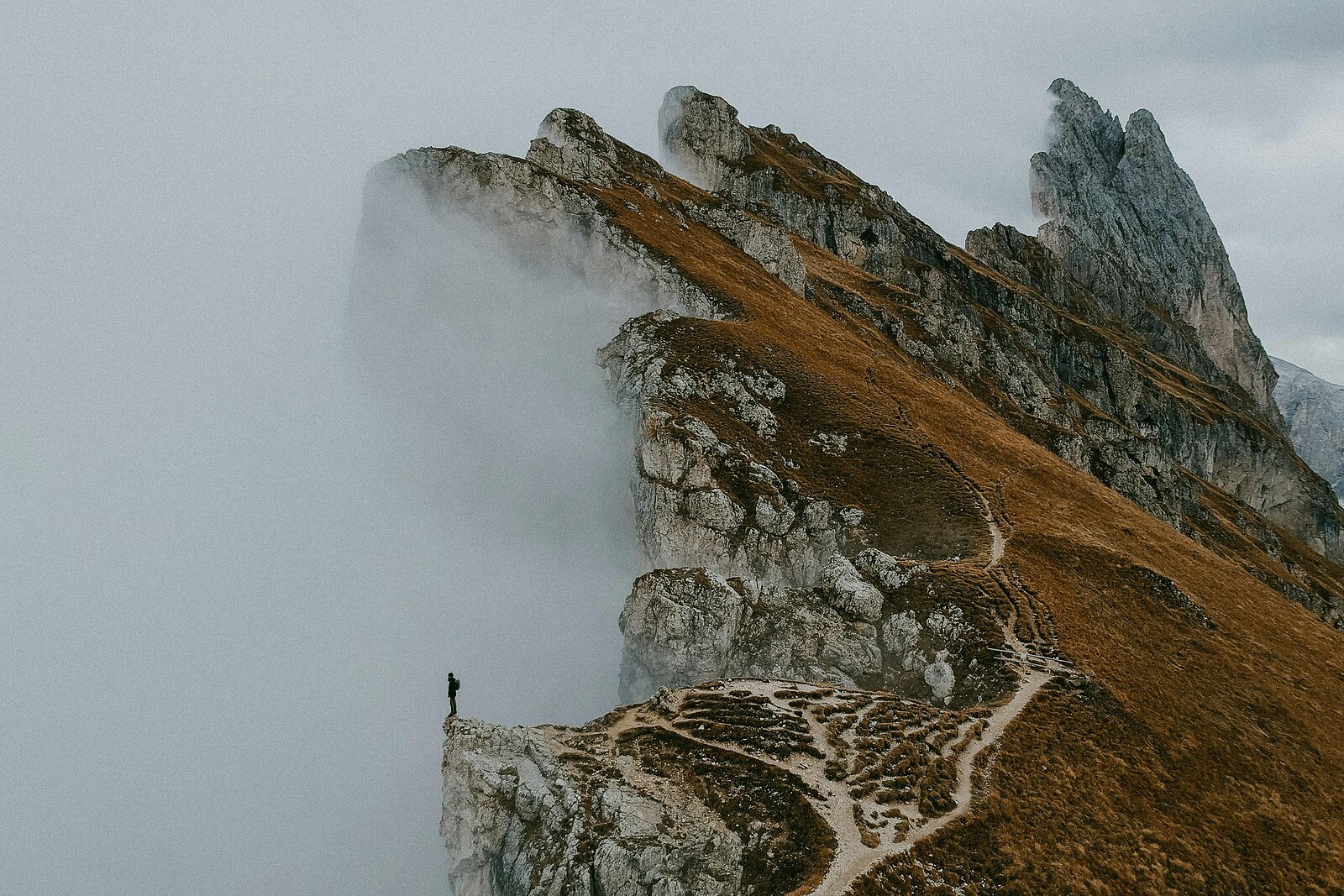 Osamělý turista stojící na skalním útesu před mlžnými horskými štíty a klikatou stezkou.